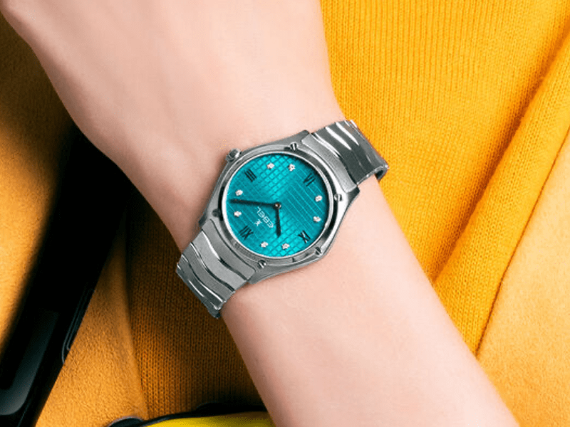 Juwelier-Wagner-Madler-Uhren-Ebel-Ansicht-Sport Eine Person trägt eine silberne Ebel-Armbanduhr mit türkisfarbenem Zifferblatt mit römischen Ziffern und kleinen dekorativen Elementen auf dem Zifferblatt. Die Person trägt ein gelbes Hemd und nur Unterarm und Hand sind sichtbar.