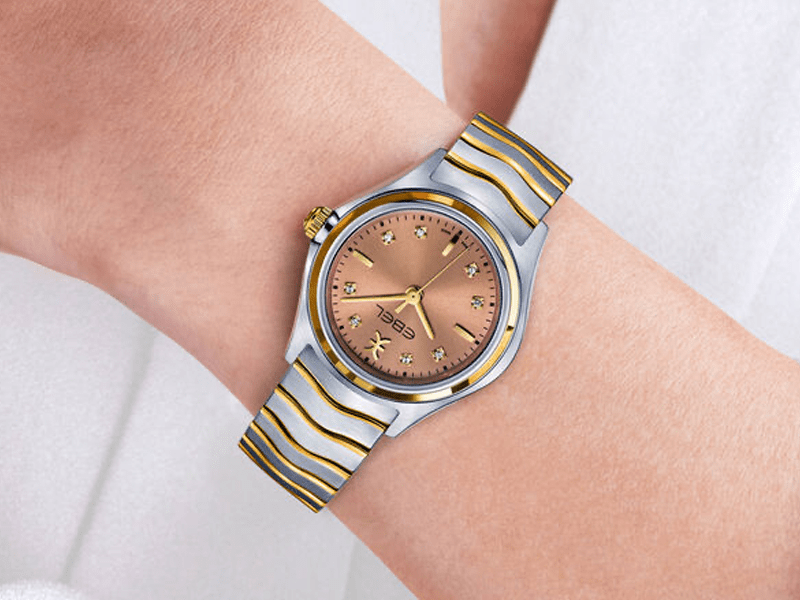 Juwelier-Wagner-Madler-Uhren-Ebel-Ansicht Eine Nahaufnahme einer Person, die eine stilvolle Ebel-Armbanduhr trägt. Die Uhr verfügt über ein rundes kupferfarbenes Zifferblatt mit goldenem Sonnenschliff, goldene Stundenmarkierungen und ein Metallarmband mit abwechselnd goldenen und silbernen Gliedern. Der Hintergrund scheint ein heller Stoff zu sein.