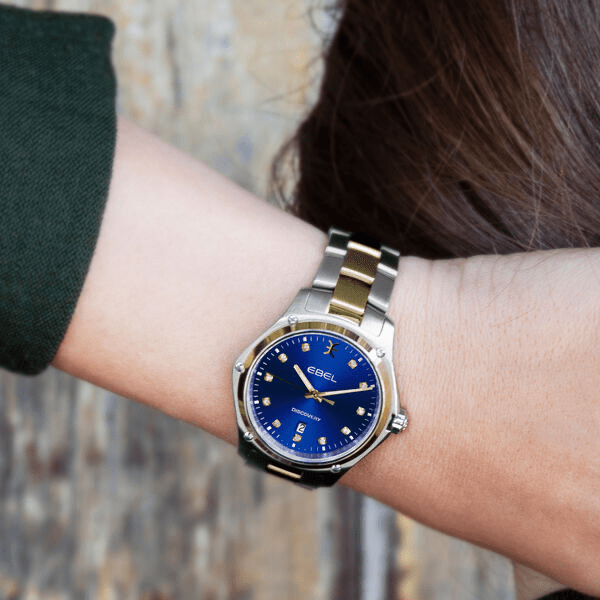 Eine Nahaufnahme eines Handgelenks, an dem eine Armbanduhr der Ebel Discovery Kollektion mit einem silber-goldenen Armband getragen wird. Die Uhr hat ein blaues Zifferblatt, goldene Stundenmarkierungen und Zeiger. Der Hintergrund scheint eine strukturierte Holzoberfläche zu sein. Die Person trägt einen grünen Ärmel.