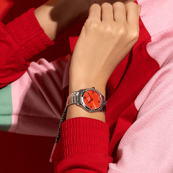 Juwelier-Wagner-Madler-Uhren-Ebel-Kollektionen-Sport Classic 1216601 Eine Person in einer schicken rot-rosa Jacke präsentiert eine Armbanduhr der Ebel Sport Classic Kollektion mit rotem Zifferblatt und silbernem Armband. Der Hintergrund ist überwiegend rot und ergänzt die Farben der Kleidung und der Uhr.