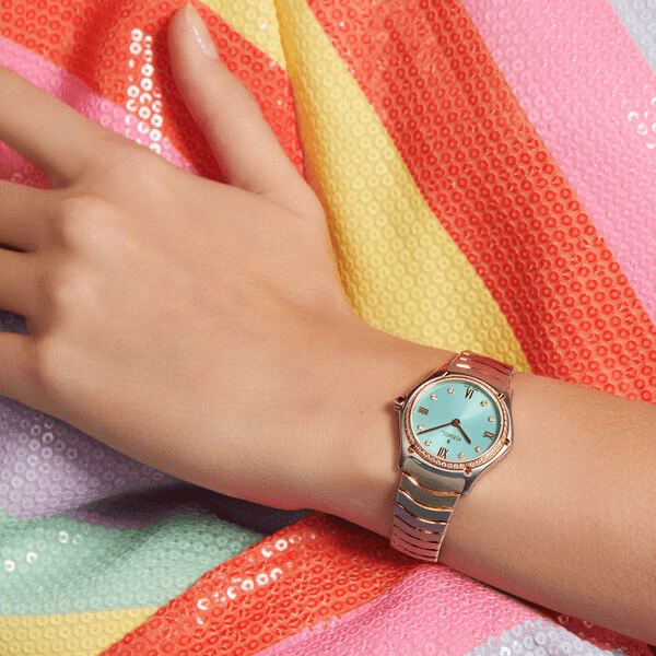 Juwelier-Wagner-Madler-Uhren-Ebel-Kollektionen-SportClassic 1216559 Eine Nahaufnahme des Handgelenks einer Person, die eine elegante goldene Armbanduhr von Ebel mit türkisem Ziffernblatt trägt. Das Handgelenk ruht auf einem farbenfrohen, mit Pailletten besetzten Stoff mit einem geometrischen Muster in Rosa, Gelb und Grün. Die Hand ist entspannt, die Finger sind leicht gespreizt.