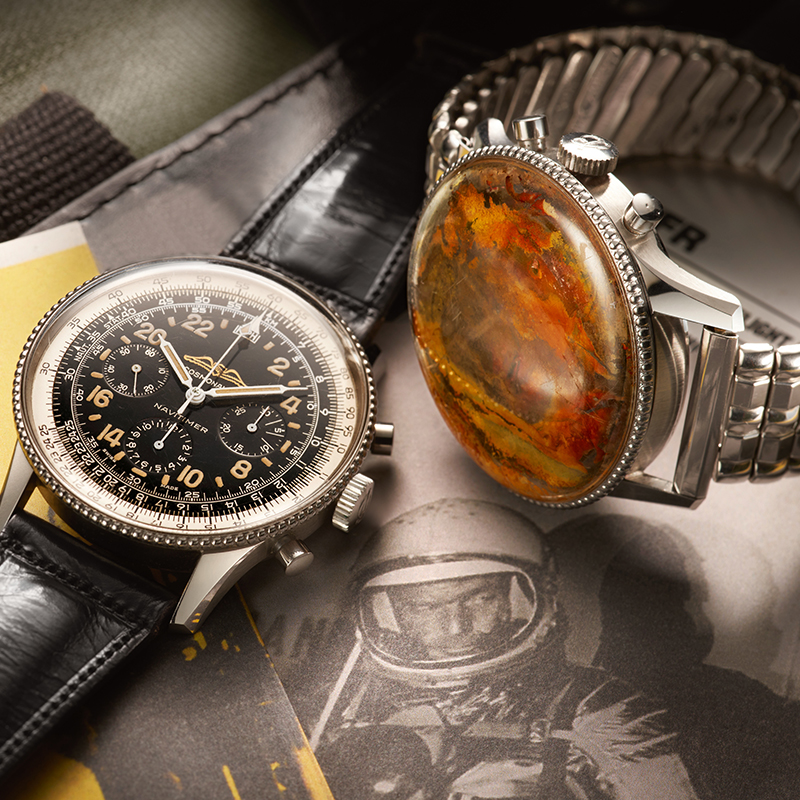 Juwelier-Wagner-Madler-Breitling-Kollektionen-Navitimer-Vorschau Auf einem Buch mit einem Astronautenbild sind zwei Armbanduhren abgebildet. Die Breitling-Uhr links hat ein schwarzes Lederarmband und ein schwarzes Zifferblatt mit mehreren Hilfszifferblättern und Markierungen. Die Uhr rechts hat ein Metallarmband und eine große, bunte, steinähnliche Abdeckung.