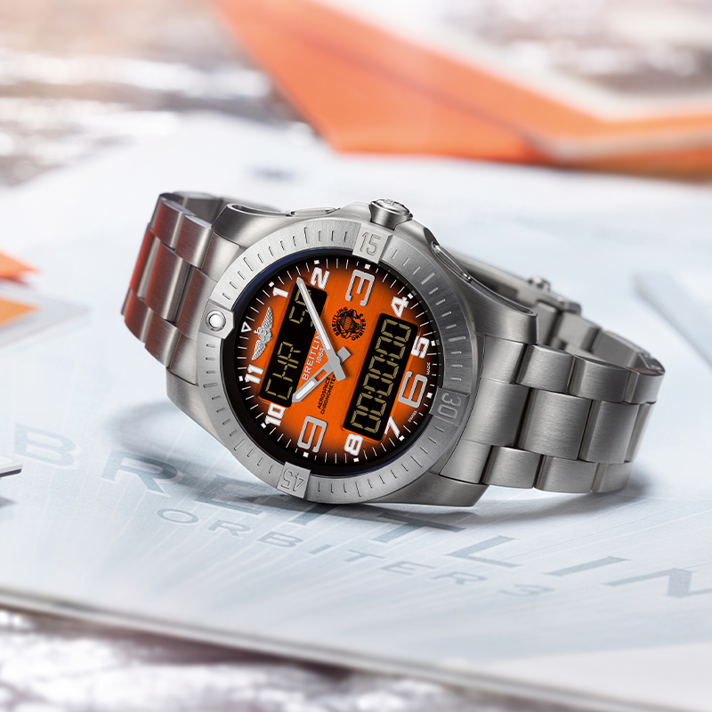 Juwelier-Wagner-Madler-Breitling-Kollektionen-Professional-Vorschau Eine Breitling-Armbanduhr aus Edelstahl mit orange-schwarzem Zifferblatt liegt auf einer Oberfläche mit bedrucktem Material. Das Zifferblatt verfügt über eine analoge Anzeige mit weißen Ziffern und digitalen Elementen, ergänzt durch ein robustes Metallgliederband.