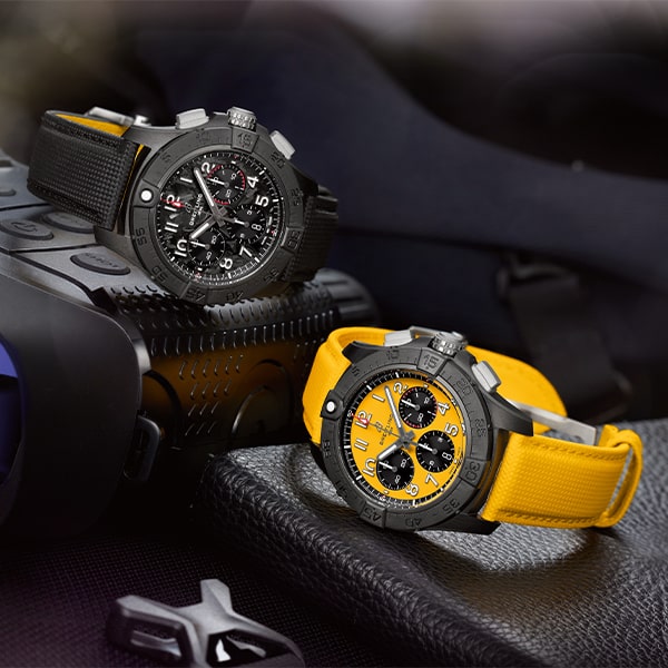 Zwei hochwertige Armbanduhren aus der Breitling Avenger Kollektion werden mit präzisen Details präsentiert. Die linke hat ein schwarzes Armband und ein schwarzes Zifferblatt mit roten Akzenten, während die rechte ein leuchtend gelbes Armband und ein gelbes Zifferblatt mit schwarzen Akzenten aufweist. Sie ruhen auf einer strukturierten Oberfläche.