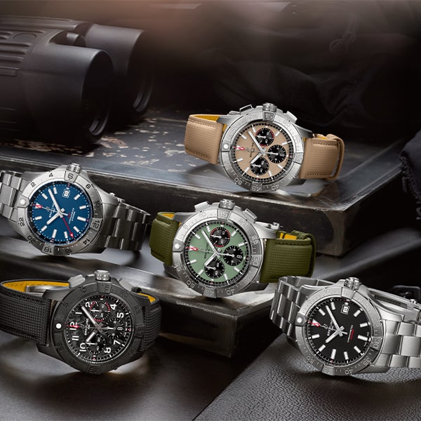 Eine Ausstellung von fünf Luxusarmbanduhren aus der Breitling Avenger Kollektion, jede mit einer metallischen Lünette und einer einzigartigen Armbandfarbe. Die Uhren sind über einem Vintage-Buch angeordnet, mit einem Fernglas im Hintergrund. Die Zifferblattfarben umfassen Blau, Schwarz, Grün und Grau, jede mit detaillierten Chronographenfunktionen.
