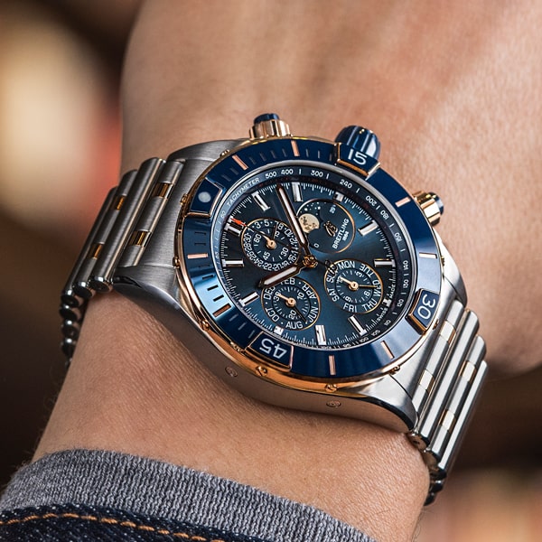 Juwelier-Wagner-Madler-Uhren-Breitling-Kollektion-Chronomat-Ansicht Eine Nahaufnahme eines Handgelenks, an dem eine luxuriöse Breitling Chronomat Kollektion Chronographenuhr mit einem silber- und goldfarbenen Metallarmband getragen wird. Das Zifferblatt ist blau mit mehreren Hilfszifferblättern und goldfarbenen Details. Die Person ist leger gekleidet, trägt graue Ärmel und eine Jeansjacke.