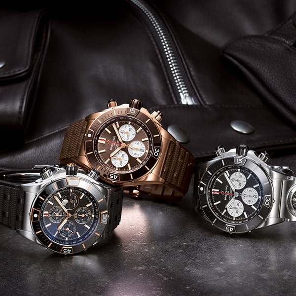 Juwelier-Wagner-Madler-Uhren-Breitling-Kollektion-Chronomat Drei luxuriöse Chronographen aus der Breitling Chronomat Kollektion werden auf einer reflektierenden Oberfläche vor dem Hintergrund einer schwarzen Lederjacke präsentiert. Die Uhren verfügen über komplizierte Zifferblätter mit mehreren Hilfszifferblättern und sind in den Ausführungen Silber, Bronze und Schwarz erhältlich, jeweils mit Gummibändern.