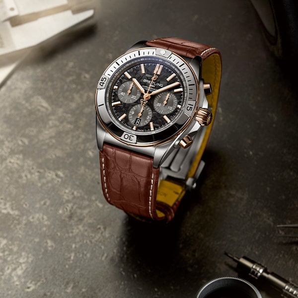 Juwelier-Wagner-Madler-Uhren-Breitling-Kollektion-Chronomat_TB01341A1B1P1 Eine stilvolle Armbanduhr mit schwarzem Zifferblatt mit mehreren Hilfszifferblättern, silbernen Akzenten und einem braunen Lederarmband. Als Teil der Breitling Chronomat Kollektion steht die Uhr auf einer flachen Oberfläche mit Uhrmacherwerkzeugen im Hintergrund, was den Eindruck einer raffinierten, hochwertigen Uhr erweckt.