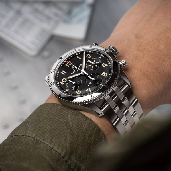Nahaufnahme des Handgelenks einer Person, die einen luxuriösen Chronographen der Breitling Classic Avi Kollektion mit schwarzem Zifferblatt, arabischen Ziffern und mehreren Hilfszifferblättern trägt. Die Person trägt eine grüne Jacke. Dokumente oder Papiere sind im Hintergrund leicht verschwommen.