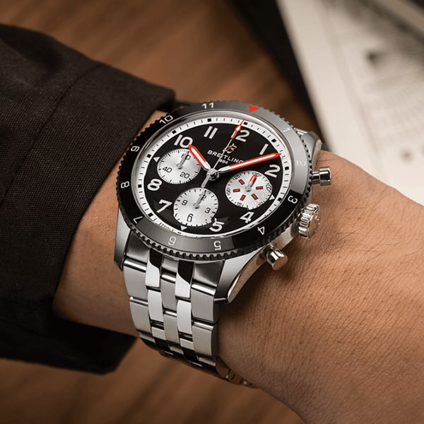 Nahaufnahme eines Handgelenks mit einer Breitling Classic Avi Kollektion, einem silbernen Chronographen mit schwarzer Lünette und weißen Zifferblättern. Die Uhr hat rote Sekundenzeiger und ein Gliederarmband aus Edelstahl. Im Hintergrund sind verschwommene Papiere zu sehen.