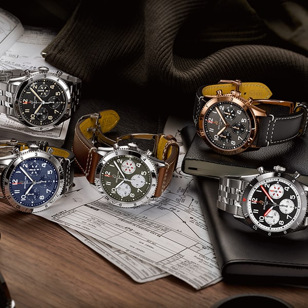 Eine Sammlung von fünf Luxusuhren aus der Breitling Classic Avi Kollektion wird auf einer Holzoberfläche mit Luftfahrtkarten und einem dunklen Stoff darunter präsentiert. Jede Uhr verfügt über ein einzigartiges Design und Farbschema, mit Metall- und Lederarmbändern, detaillierten Zifferblättern und verschiedenen Chronographenfunktionen.