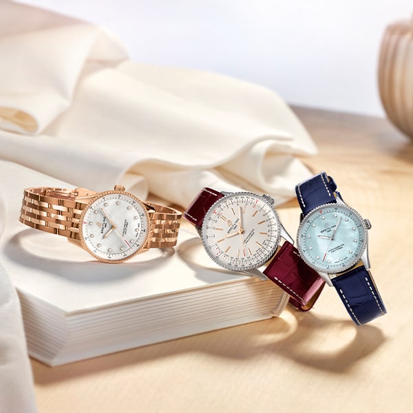 Drei elegante Armbanduhren aus der Breitling Navitimer Kollektion werden auf einer hellen Holzoberfläche mit weißem Stoffhintergrund präsentiert. Eine Uhr hat ein Armband aus Roségold, eine andere ein kastanienbraunes Lederarmband und die dritte ein marineblaues Lederarmband. Alle sind mit funkelnden Details auf ihren Zifferblättern geschmückt.