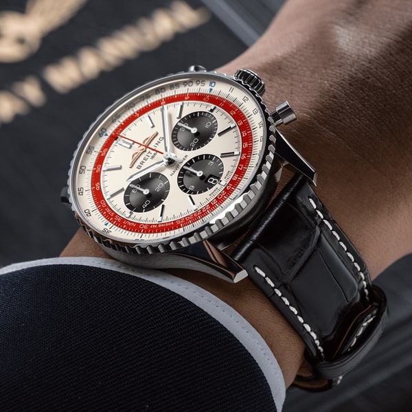 Nahaufnahme einer Person, die eine Luxusarmbanduhr aus der Breitling Navitimer Kollektion trägt. Die Uhr hat ein rundes silbernes Gehäuse, ein weißes Zifferblatt mit schwarzen Hilfszifferblättern und einen roten Akzentring um die Mitte. Sie hat ein schwarzes Lederarmband mit weißen Nähten. Die Person trägt einen dunklen Anzug.