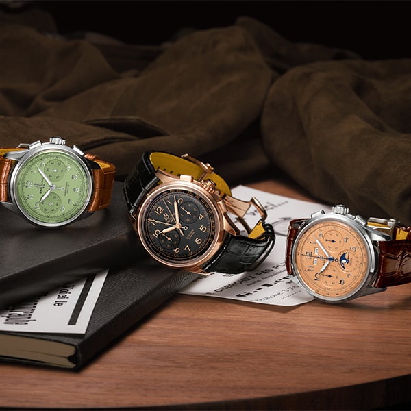 Drei Luxusarmbanduhren aus der Breitling Premier Kollektion mit Lederarmbändern werden auf einer Holzoberfläche präsentiert. Die Uhren haben unterschiedliche Zifferblattfarben: Grün, Schwarz und Lachs. Dahinter befinden sich dunkle Stoffstücke, ein Dokument und gestapelte Bücher. Die Inszenierung ist elegant und raffiniert.