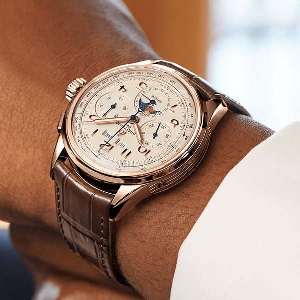 Eine Nahaufnahme des Handgelenks einer Person, die eine luxuriöse Breitling Premier Kollektion-Armbanduhr mit braunem Lederarmband und detailreichem weißen Zifferblatt trägt. Die Uhr verfügt über mehrere Hilfszifferblätter, darunter Mondphasen und verschiedene andere Komplikationen, eingefasst in einen Rahmen aus Gold oder Roségold.