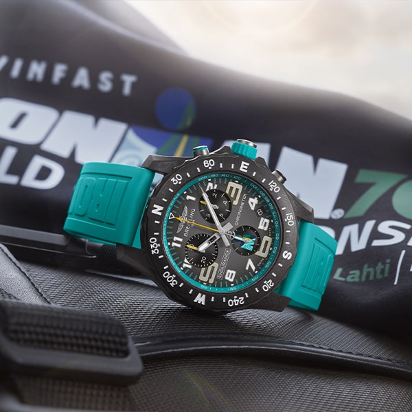 Eine stilvolle Armbanduhr aus der Breitling Professional Kollektion mit türkisem Armband und dunklem Ziffernblatt, das die Uhrzeit und verschiedene Zifferblätter anzeigt, liegt auf einer Fläche. Im Hintergrund ist ein Kleidungsstück oder Stoffstück mit weißem und blauem Text teilweise sichtbar.