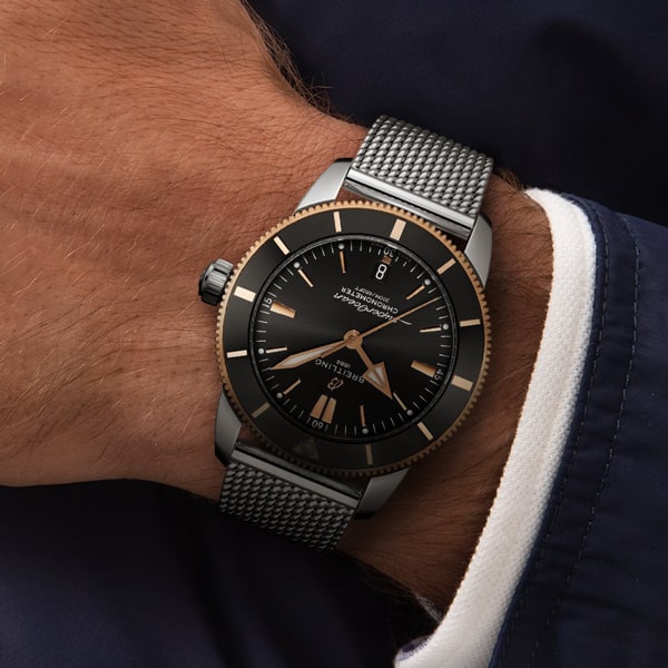 Juwelier-Wagner-Madler-Uhren-Breitling-Kollektion-Superocean-Heritage-Ansicht Eine Nahaufnahme des Handgelenks einer Person, die eine stilvolle Breitling Super Ocean Heritage-Uhr mit schwarzem Zifferblatt, roségoldenen Markierungen und einem Metallgeflechtarmband trägt. Die Person trägt eine dunkelblaue Jacke mit einer weißen Manschette. Auf dem Zifferblatt der Uhr sind der Markenname und andere Details zu sehen.