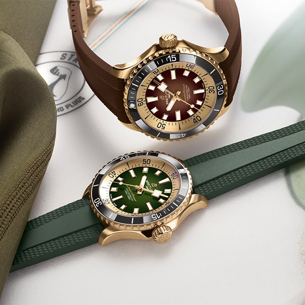 Es werden zwei Armbanduhren aus der Breitling Superocean Kollektion mit Taucherlünette und goldfarbenen Gehäusen ausgestellt. Die obere Uhr hat ein braunes Armband und ein rotes Zifferblatt, während die untere Uhr ein grünes Armband und ein grünes Zifferblatt hat. Beide Uhren liegen auf einer hellen Oberfläche zwischen grünem und braunem Stoff.