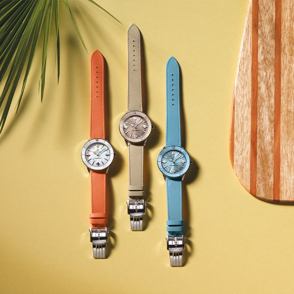 Juwelier-Wagner-Madler-Uhren-Breitling-Kollektion-Superocean_Heritage Drei Breitling Super Ocean Heritage-Armbanduhren sind vertikal auf einem gelben Hintergrund angeordnet, jede mit Lederarmbändern in Orange, Beige und Blau. Die Zifferblätter sind silberfarben mit minimalistischem Design. Ein grünes Palmblatt wirft einen Schatten aus der oberen linken Ecke und ein Holzpaddel befindet sich auf der rechten Seite.