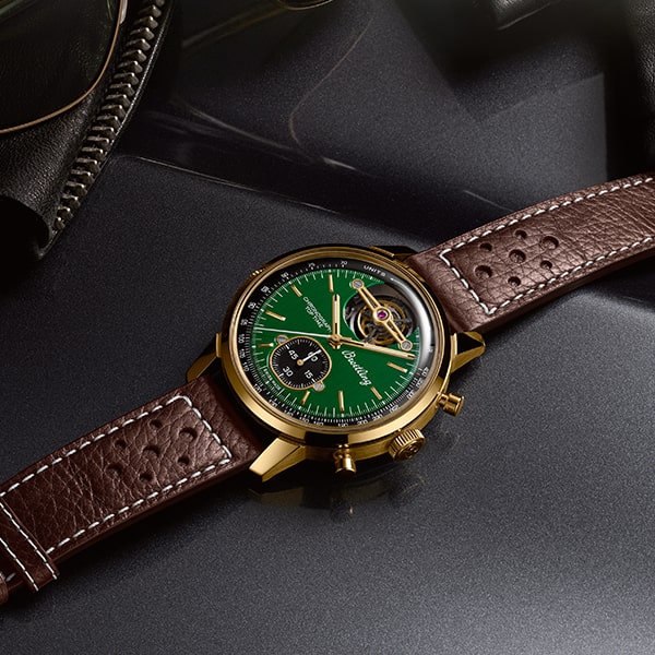 Eine stilvolle Armbanduhr aus der Breitling Top Time Kollektion mit einem grün-goldenen Zifferblatt und einem braunen Lederarmband mit weißen Nähten liegt auf einer reflektierenden dunklen Oberfläche. Die Uhr verfügt über eine Chronographenfunktion und sichtbare Zahnräder sowie ein schwarzes Hilfszifferblatt für zusätzliche Funktionalität.