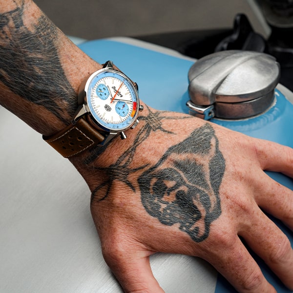 Nahaufnahme einer tätowierten Hand, die eine Armbanduhr aus der Breitling Top Time Kollektion trägt. Die Uhr hat ein braunes Lederarmband und ein farbenfrohes Zifferblatt mit blauen, weißen und orangefarbenen Akzenten. Der Zeiger ruht auf einer metallischen Oberfläche, wahrscheinlich einem Teil eines Fahrzeugs mit sichtbarem Tankdeckel.