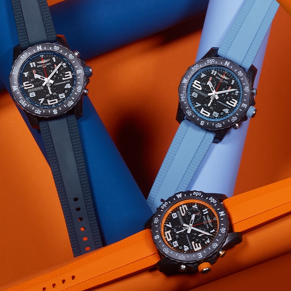 Es werden drei Fliegerarmbanduhren der Breitling Professional Kollektion mit schwarzen Zifferblättern und fettgedruckten Ziffern angezeigt. Die linke hat ein schwarzes Armband, die obere rechte hat ein blaues Armband und die untere rechte hat ein orangefarbenes Armband. Der Hintergrund weist Streifenmuster in Orange- und Blautönen auf.