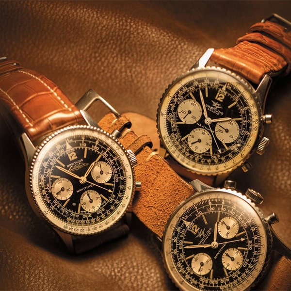 Juwelier-Wagner-Madler-Uhren-Breitling-Navitimer-Heritage Drei Vintage-Armbanduhren von Breitling mit braunen Lederarmbändern werden auf einer strukturierten braunen Oberfläche präsentiert. Die Uhren verfügen über schwarze Zifferblätter mit weißen Chronographen-Hilfszifferblättern und silbernen Einfassungen, die komplizierte Markierungen und Zeiger zeigen und eine klassische, zeitlose Ästhetik hervorrufen.