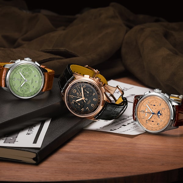 Juwelier-Wagner-Madler-Uhren-Breitling-Premier-Vorschau Drei luxuriöse Breitling-Uhren auf einer Holzoberfläche. Die erste Uhr links hat ein grünes Zifferblatt mit silberner Lünette und braunem Lederarmband. Die mittlere Uhr hat ein schwarzes Zifferblatt mit goldener Lünette und schwarzem Lederarmband. Die rechte Uhr hat ein orangefarbenes Zifferblatt mit silberner Lünette und braunem Lederarmband. Sie liegen auf einem Notizbuch und einigen Papieren, mit einem braunen Tuch im