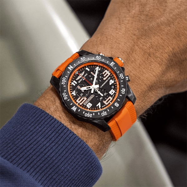 Juwelier-Wagner-Madler-Uhren-Breitling-Top-Time-X82310A51B1S2 Eine Nahaufnahme des Handgelenks einer Person, die eine schwarz-orange Breitling-Armbanduhr trägt. Die Uhr hat ein schwarzes Zifferblatt mit weißen Ziffern und orangefarbenen Akzenten. Das Armband ist orange und bildet einen perfekten Kontrast zu dem blauen Langarmshirt, das die Person trägt.