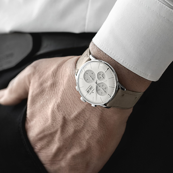 Juwelier-Wagner-Madler-Uhren–Junghans-Kollektion-Ansicht-2 Eine Nahaufnahme des Handgelenks einer Person, die eine elegante, silberne Junghans-Chronographenuhr mit einem hellen Lederarmband trägt. Die Hand der Person ruht lässig in einer schwarzen Hosentasche und sie trägt ein weißes Langarmhemd. Das Zifferblatt zeigt mehrere Hilfszifferblätter.