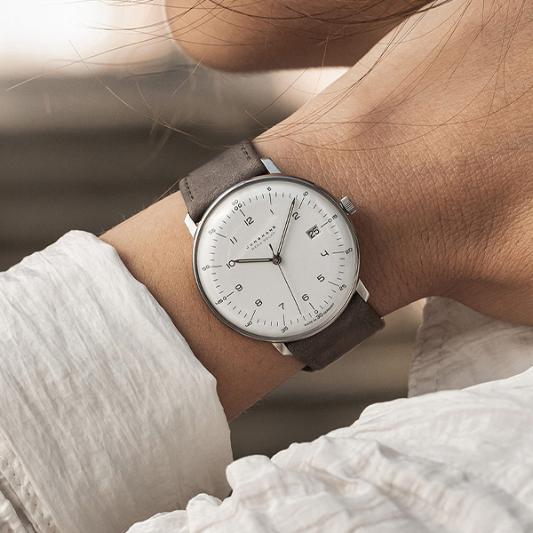 Juwelier-Wagner-Madler-Uhren–Junghans-Kollektion-Ansicht-3 Eine Nahaufnahme eines Handgelenks, an dem eine minimalistische Junghans-Uhr mit weißem Zifferblatt, dünnen schwarzen Zeigern und einem braunen Lederarmband getragen wird. Die Person trägt ein weißes Langarmhemd. Der Hintergrund ist unscharf und lenkt die Aufmerksamkeit auf die Uhr.