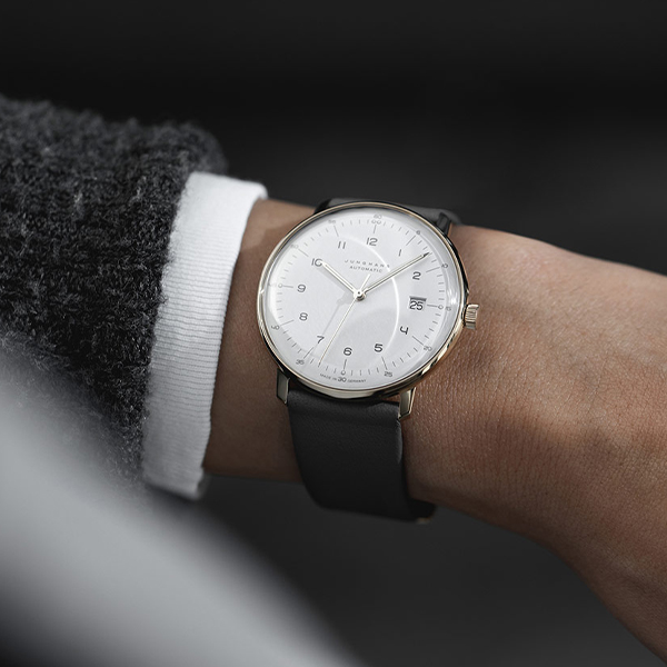 Juwelier-Wagner-Madler-Uhren–Junghans-Kollektion-Ansicht Eine Nahaufnahme einer Person, die eine Junghans-Armbanduhr mit weißem Zifferblatt, schwarzem Lederarmband und goldenem Gehäuse trägt. Die Uhr weist ein minimalistisches Design mit schwarzen Ziffern und Zeigern sowie einem Datumsfenster auf. Die Person trägt eine graue Tweedjacke mit einer weißen Hemdmanschette.