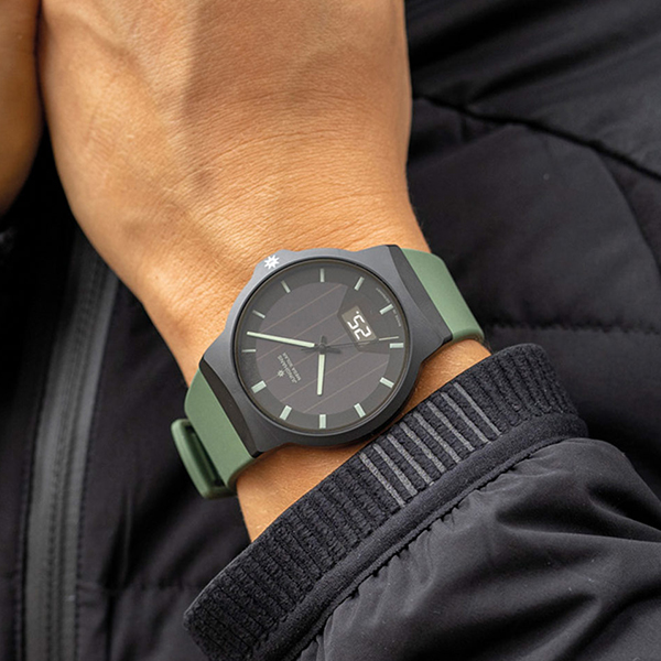 Juwelier-Wagner-Madler-Uhren-Junghans-Kollektion-force-ansicht-2 Eine Nahaufnahme des Handgelenks einer Person, die eine schwarze Uhr aus der Junghans Force Kollektion mit grünem Armband trägt. Die Uhr hat ein minimalistisches Design mit schwarzem Zifferblatt, hellgrünen Stundenmarkierungen und einer kleinen Digitalanzeige auf der rechten Seite. Die Person trägt eine schwarze Steppjacke.