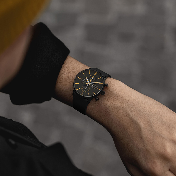 Juwelier-Wagner-Madler-Uhren-Junghans-Kollektion-form-ansicht-3 Nahaufnahme des Handgelenks einer Person, die eine stilvolle schwarze Armbanduhr mit goldenen Akzenten aus der Junghans Form Kollektion trägt. Die Uhr hat ein schlankes, modernes Design mit einem schwarzen Armband. Die Person trägt dunkle Kleidung und steht vor einem verschwommenen, rustikalen Kopfsteinpflasterhintergrund, wodurch die Aufmerksamkeit auf die Uhr gelenkt wird.