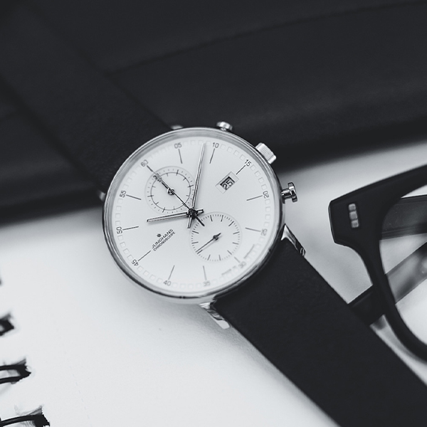 Juwelier-Wagner-Madler-Uhren-Junghans-Kollektion-form-ansicht Eine Nahaufnahme einer analogen Armbanduhr aus der Junghans Form Kollektion in Schwarzweiß mit zwei kleineren Hilfszifferblättern und einem Datumsfenster. Die Uhr mit dem schwarzen Lederarmband liegt neben einer Brille auf einer weißen Oberfläche, im Hintergrund ist teilweise ein Notizbuch zu sehen.