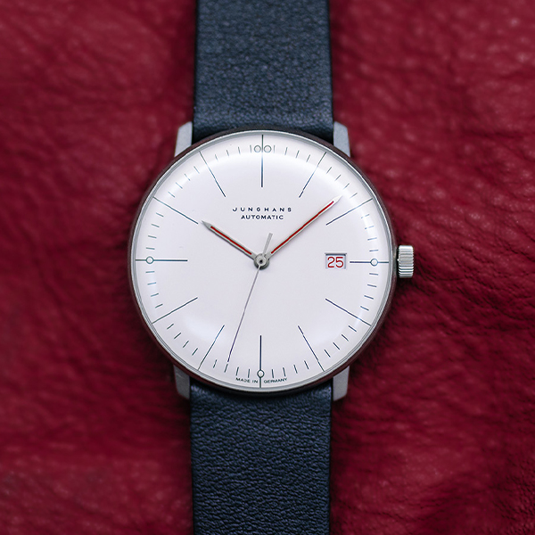Juwelier-Wagner-Madler-Uhren-Junghans-Kollektion-max-bill-ansicht-2 Eine minimalistische Junghans Max Bill Armbanduhr mit weißem Zifferblatt, schwarzem Lederarmband und silbernem Gehäuse ist vor einem strukturierten roten Hintergrund zu sehen. Sie zeigt das Datum „25“ in einem kleinen roten Kästchen. Die Uhr hat dünne schwarze Stundenmarkierungen und Zeiger sowie einen roten Sekundenzeiger. Die Marke „Junghans Automatic“ ist auf dem Zifferblatt sichtbar.