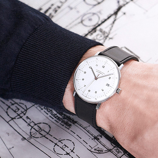 Juwelier-Wagner-Madler-Uhren-Junghans-Kollektion-max-bill-ansicht -3 Eine Person in einem dunklen Langarmhemd zeigt ihr Handgelenk mit einer minimalistischen silbernen Junghans Max Bill-Armbanduhr mit weißem Zifferblatt und schwarzem Lederarmband. Im Hintergrund sind technische Zeichnungen oder Blaupausen auf einer Oberfläche ausgebreitet.