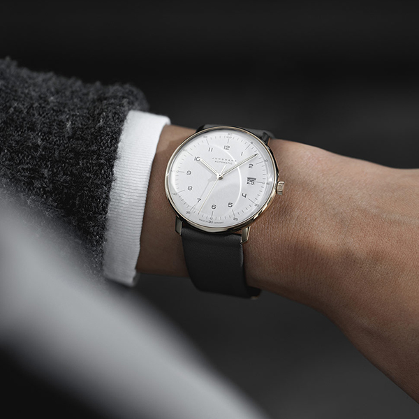 Juwelier-Wagner-Madler-Uhren-Junghans-Kollektion-max-bill-ansicht Nahaufnahme des Handgelenks einer Person, die eine elegante, minimalistische silberne Junghans Max Bill-Armbanduhr mit schwarzem Lederarmband trägt. Die Uhr hat ein weißes Zifferblatt mit schwarzen Stundenmarkierungen und einem kleinen Datumsfenster. Die Person trägt einen dunklen Anzug mit weißem Hemd.