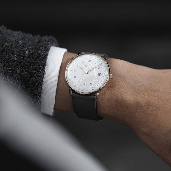Juwelier-Wagner-Madler-Uhren-Junghans-Kollektion-max-bill-automatic-ansicht-2 Eine Nahaufnahme des Handgelenks einer Person, die eine stilvolle Armbanduhr aus der Junghans Max Bill Automatic Kollektion trägt, mit weißem Zifferblatt, silbernem Gehäuse und schwarzem Lederarmband. Die Person trägt einen dunklen Ärmel mit einer weißen Manschette. Der unscharfe Hintergrund lenkt die Aufmerksamkeit auf die Uhr.