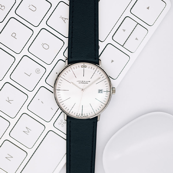 Juwelier-Wagner-Madler-Uhren-Junghans-Kollektion-max-bill-automatic-ansicht Eine elegante Armbanduhr der Junghans Max Bill Automatic Kollektion mit schwarzem Lederarmband und weißem Zifferblatt mit Datumsanzeige und dünnen schwarzen Zeigern steht auf einer weißen Computertastatur. Rechts neben der Uhr ist teilweise eine weiße Computermaus zu sehen.