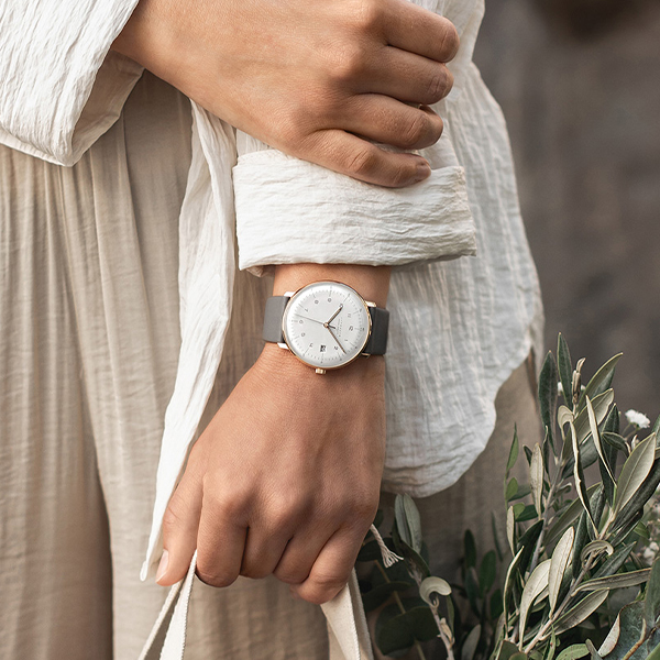 Juwelier-Wagner-Madler-Uhren-Junghans-Kollektion-max-bill-funk-and-solar-ansicht Eine Person mit heller Hautfarbe trägt ein lockeres, helles Langarmhemd und eine Armbanduhr der Junghans Max Bill Funk & Solar Kollektion mit weißem Zifferblatt und grauem Armband. Sie hält eine Einkaufstasche und eine grüne Pflanze in einer Hand, vor einem unscharfen Hintergrund.