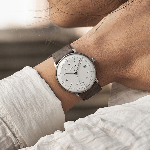Juwelier-Wagner-Madler-Uhren-Junghans-Kollektion-max-bill-funk-und-solar-ansicht-2 Eine Nahaufnahme einer Person, die eine minimalistische Armbanduhr der Junghans Max Bill Funk & Solar Kollektion mit weißem Zifferblatt und grauem Lederarmband trägt. Das schlichte Design der Uhr zeichnet sich durch dünne Stunden- und Minutenzeiger, kleine Ziffern und ein schlankes Edelstahlgehäuse aus. Die Person trägt ein weißes Langarmhemd.