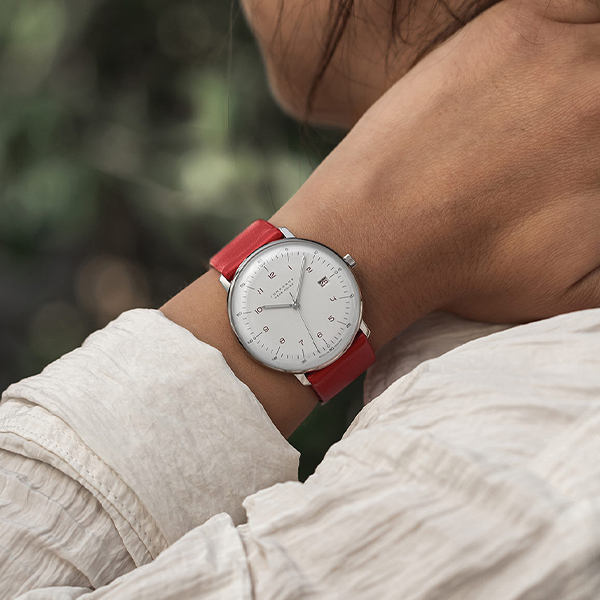 Juwelier-Wagner-Madler-Uhren-Junghans-Kollektion-max-bill-funk-und-solar-ansicht-3 Eine Person mit heller Hautfarbe und weißem Hemd hält sich mit der linken Hand den Hals und präsentiert eine Uhr der Junghans Max Bill Funk & Solar Kollektion mit weißem Zifferblatt und rotem Lederarmband. Der Hintergrund ist leicht verschwommen mit Grüntönen.