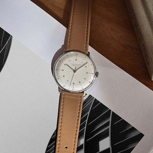 Juwelier-Wagner-Madler-Uhren-Junghans-Kollektion-max-bill-handaufzug-ansicht-2 Eine minimalistische Armbanduhr aus der Junghans Max Bill Kollektion mit hellbraunem Lederarmband und weißem Zifferblatt mit schlichten schwarzen Stundenmarkierungen und Zeigern ruht auf einem Blatt Papier mit geometrischen schwarz-weißen Mustern. Die Szene spielt auf einer Holzoberfläche, an deren Rand ein Buch zu sehen ist.