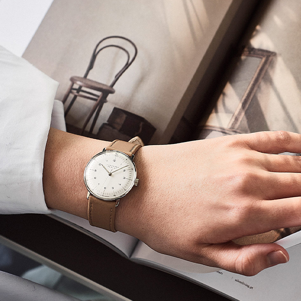 Juwelier-Wagner-Madler-Uhren-Junghans-Kollektion-max-bill-handaufzug-ansicht-3 Eine Person trägt eine elegante, minimalistische Junghans Max Bill Hansaufzug-Armbanduhr mit hellbraunem Lederarmband und weißem Zifferblatt und legt ihre Hand auf ein offenes Buch. Im Hintergrund sind ein verschwommenes Bild eines Holzstuhls und gerahmte Bilder zu sehen, was der Szene eine Vintage-Atmosphäre verleiht.