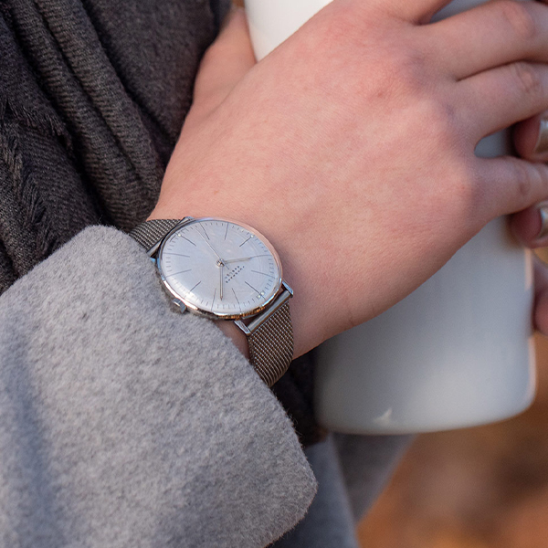 Juwelier-Wagner-Madler-Uhren-Junghans-Kollektion-max-bill-handaufzug-ansicht Nahaufnahme der Hand einer Person, die einen grauen Pullover trägt und einen weißen Becher hält. Die Person trägt außerdem eine silberne Junghans Max Bill Hansaufzug-Armbanduhr mit Mesh-Armband, die prominent am Handgelenk zu sehen ist. Der Hintergrund ist verschwommen, sodass die Aufmerksamkeit auf die Hand, den Becher und die Uhr gerichtet ist.
