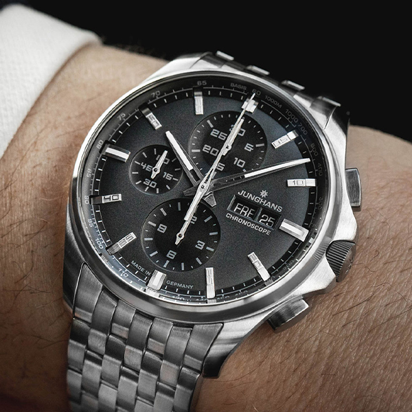 Juwelier-Wagner-Madler-Uhren-Junghans-Kollektion-meister-ansicht-2 Eine Nahaufnahme eines Handgelenks, an dem eine silberne Junghans Chronoscope-Uhr aus der angesehenen Junghans Meister Kollektion getragen wird. Die Uhr verfügt über ein schwarzes Zifferblatt mit drei Hilfszifferblättern, silbernen Stundenmarkierungen, Datums- und Tagesanzeige und silbernen Zeigern, alles umhüllt von einem Edelstahlarmband mit mehreren Gliedern.