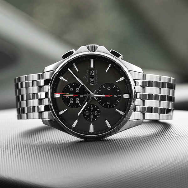 Juwelier-Wagner-Madler-Uhren-Junghans-Kollektion-meister-ansicht-3 Eine stilvolle analoge Armbanduhr aus der Junghans Meister Kollektion mit schwarzem Zifferblatt und silbernem Metallarmband liegt auf einer strukturierten Oberfläche. Die Uhr verfügt über drei Hilfszifferblätter, eine Tages- und Datumsanzeige sowie leuchtende Zeiger und Markierungen und strahlt ein schlankes und anspruchsvolles Design aus.