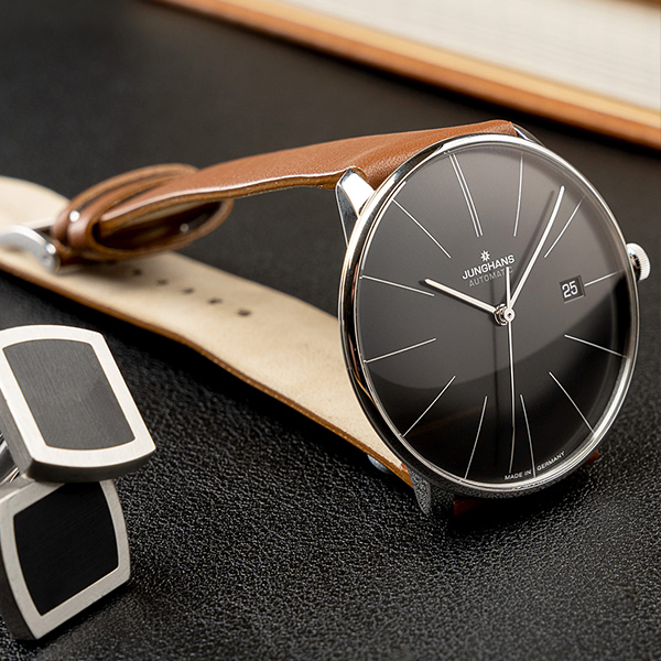 Juwelier-Wagner-Madler-Uhren-Junghans-Kollektion-meister-ansicht Eine minimalistische Armbanduhr der Junghans Meister Kollektion mit schwarzem Zifferblatt, schmalen silbernen Stundenmarkierungen und Zeigern und einem braunen Lederarmband liegt auf einer dunklen Lederoberfläche. Die Uhr zeigt das Datum auf der 3-Uhr-Position. Daneben befindet sich ein Paar silberne und schwarze Manschettenknöpfe.