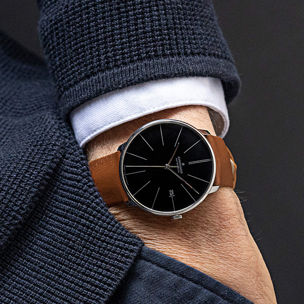 Juwelier-Wagner-Madler-Uhren-Junghans-Kollektion-meister-automatic-ansicht-2 Nahaufnahme des Handgelenks einer Person, die eine elegante Armbanduhr der Junghans Meister Automatik Kollektion trägt. Die Uhr hat ein elegantes schwarzes Zifferblatt mit minimalistischem Design, silberne Stundenmarkierungen und ein braunes Lederarmband. Die Person trägt eine strukturierte marineblaue Jacke und ein weißes Oberhemd.